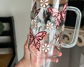 Puede incluir: Un vaso de vidrio con tapa de bambú y una pajita. El vaso tiene un diseño de mariposas rosas y marrones con flores blancas.