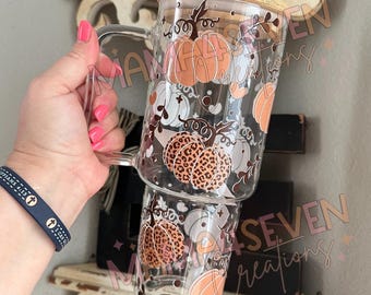 May include: Clear glass mug with a handle, decorated with orange, white, and leopard print pumpkins. The mug has a wooden lid and is designed for fall. The mug is held up in the air.