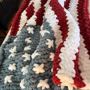 May include: A handmade, patriotic blanket featuring a design inspired by the American flag. The blanket has alternating red and white stripes, with a section of blue fabric adorned with white star-shaped accents. The chunky knit construction adds texture.