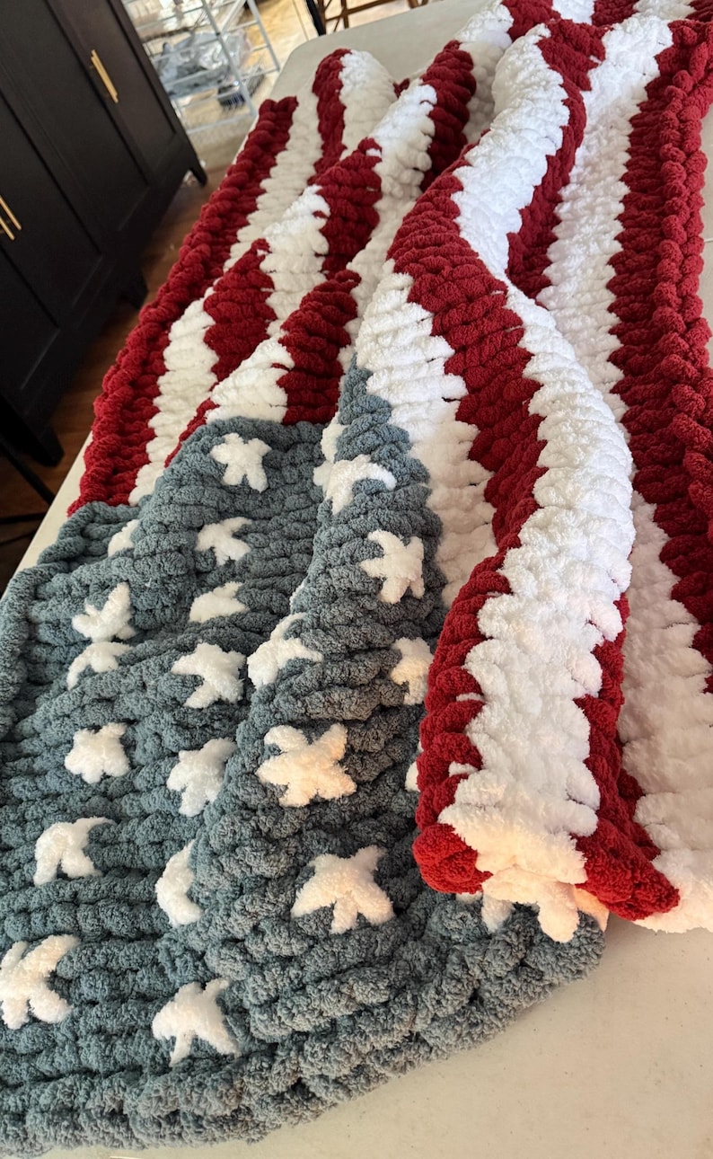 May include: A handmade, patriotic blanket featuring a design inspired by the American flag. The blanket has alternating red and white stripes, with a section of blue fabric adorned with white star-shaped accents. The chunky knit construction adds texture.