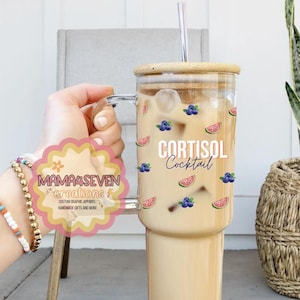 May include: A clear glass tumbler with a wooden lid and a straw. The tumbler is filled with a light brown liquid and ice. The tumbler has a design of watermelons and blueberries. The text "CORTISOL Cocktail" is printed on the tumbler.