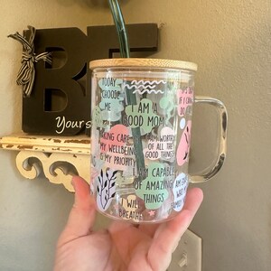 May include: A clear glass mug with a bamboo lid and a green straw. The mug has various colorful stickers with positive affirmations, including "I am a good mom", "I will breathe", and "I am capable of amazing things".