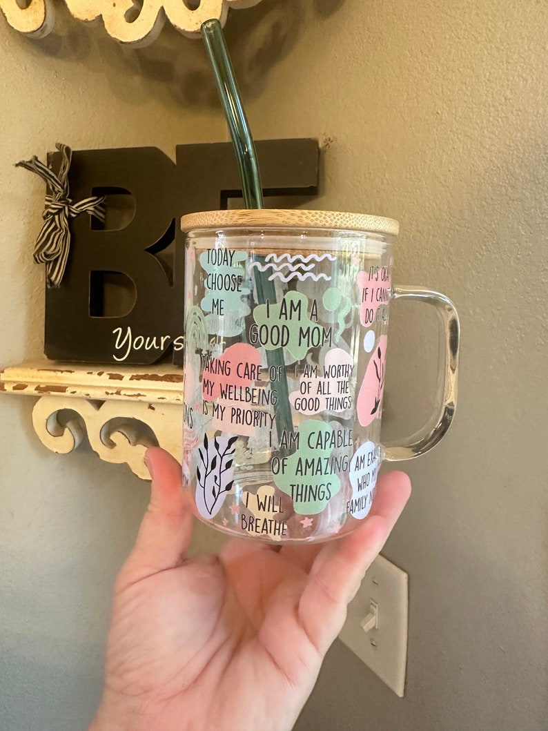May include: A clear glass mug with a bamboo lid and a green straw. The mug has various colorful stickers with positive affirmations, including "I am a good mom", "I will breathe", and "I am capable of amazing things".