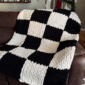 May include: A chunky knit blanket in a black and white checkerboard pattern. The blanket is draped over a brown leather couch. The large squares of the blanket are approximately 12 inches by 12 inches.