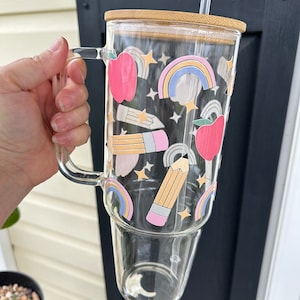 May include: A clear glass tumbler with a bamboo lid and a straw. The tumbler has a colorful design with rainbows, stars, pencils, and apples.