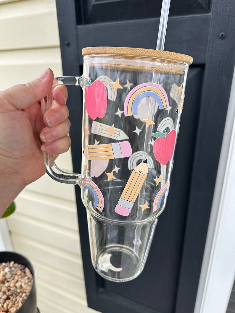 May include: A clear glass tumbler with a bamboo lid and a straw. The tumbler has a colorful design with rainbows, stars, pencils, and apples.