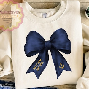 May include: Cream-colored sweatshirt featuring a large navy blue bow graphic. The bow has a gold anchor and the text "NAVY MOM EST. 2025". The sweatshirt is a crew neck style.