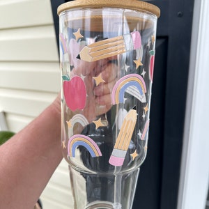 May include: A clear glass tumbler with a bamboo lid and a straw. The tumbler has a colorful design with rainbows, stars, pencils, and apples.