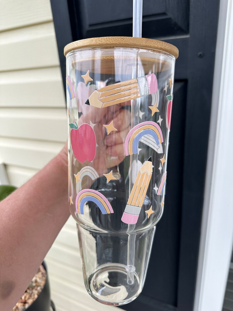 May include: A clear glass tumbler with a bamboo lid and a straw. The tumbler has a colorful design with rainbows, stars, pencils, and apples.