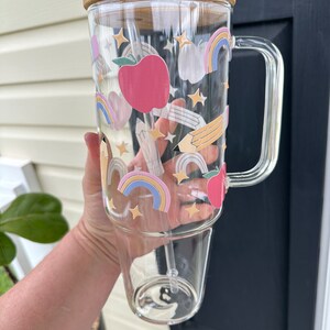 May include: A clear glass tumbler with a bamboo lid and a straw. The tumbler has a colorful design with rainbows, stars, pencils, and apples.