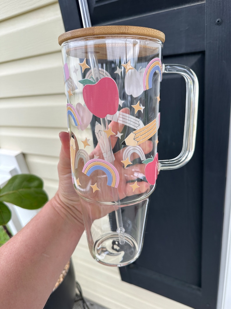 May include: A clear glass tumbler with a bamboo lid and a straw. The tumbler has a colorful design with rainbows, stars, pencils, and apples.