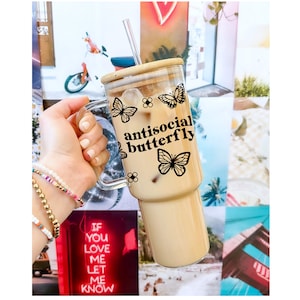 May include: A clear glass tumbler with a handle, filled with iced coffee. The tumbler features a bamboo lid, a reusable straw, and a black butterfly design with the words "antisocial butterfly". The background is a collage of colorful images.