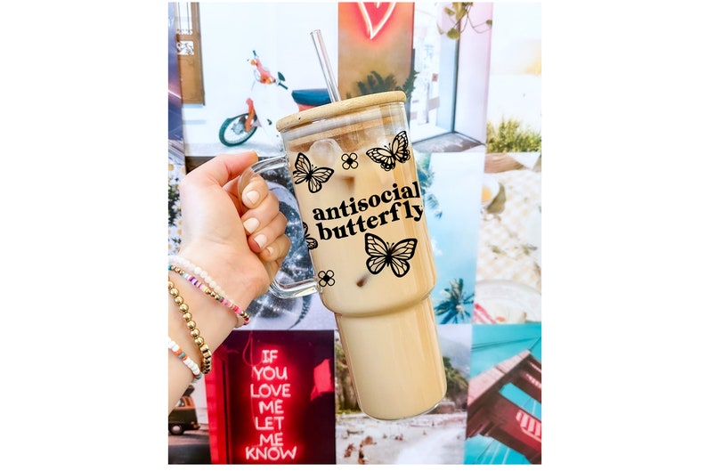 May include: A clear glass tumbler with a handle, filled with iced coffee. The tumbler features a bamboo lid, a reusable straw, and a black butterfly design with the words "antisocial butterfly". The background is a collage of colorful images.