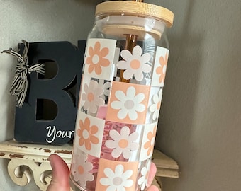 Puede incluir: Una botella de agua reutilizable de vidrio transparente con tapa de bambú y una pajita. La botella tiene un patrón de margaritas rosas y blancas.