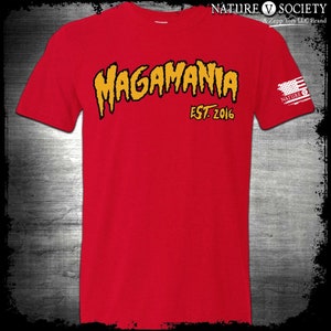 以下が含まれることがあります： 「MAGAMANIA EST. 2016」と書いてある黄色と黒のグラフィックがプリントされた赤いTシャツ。右袖には小さなアメリカ国旗のパッチがあり、その上に「NATURE V SOCIETY」の文字が書かれている。