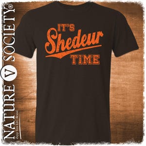 Peut inclure: Un t-shirt marron foncé avec le texte orange "IT'S Shedeur TIME". Le texte a un aspect vieilli et vintage. Le t-shirt est de style col rond.