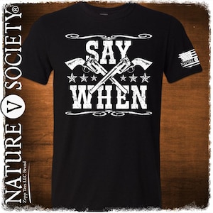 May include: Black t-shirt with a white graphic design that says "SAY WHEN" with two crossed pistols and stars. The graphic is in a vintage style. The shirt has a small logo on the right sleeve that says "NATURE V SOCIETY".
