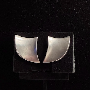 May include: A pair of silver-toned earrings with a smooth, curved, fan-like shape. Each earring features a concave edge and a polished metallic finish, presenting a sleek, geometric design that tapers toward the bottom in a symmetrical arrangement.