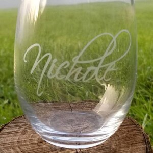 May include: A clear glass wine glass with the name "Nicholas" etched into the side. The glass is sitting on a wooden surface.
