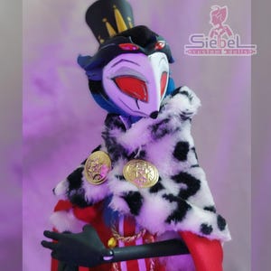 Op de afbeelding: Een pluche pop van een personage met een wit masker, rode ogen en blauw haar, gekleed in een zwarte hoge hoed, een zwart-witte gevlekte bontkraag en een rode jas met gouden accenten. De pop heeft zwarte handschoenen en een gouden ketting.
