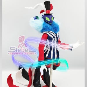 May include: A custom doll with a theatrical design. The doll features a top hat, a blue and white striped vest, and red sleeves. The doll has a blue and white furry collar and a purple mask with green eyes. The doll's name is Siebel Custom Dolls.