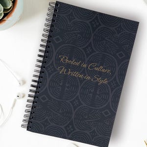 May include: A spiral-bound notebook with a dark blue cover featuring a repeating circular pattern. The cover has the text "Rooted in Culture, Written in Style" in gold lettering. The notebook is open, revealing the spiral binding and blank pages.