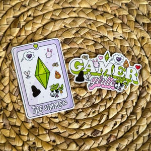 May include: Two stickers on a woven background. One sticker is a rectangular card with a green diamond and the text "THE SIMMER". The other sticker says "GAMER girlie" in green and pink lettering, with various graphics.