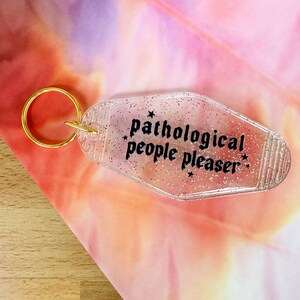 May include: Clear acrylic keychain with the text "pathological people pleaser" in black. The keychain has a gold-colored ring and is decorated with glitter and small star shapes. The background is a blend of pink, orange, and purple.