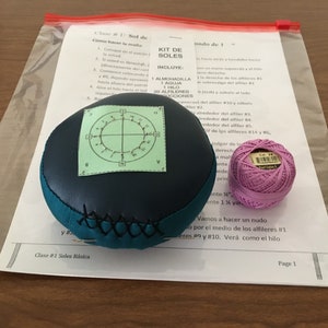 May include: A kit for making soles, including a dark blue and teal cushion with a green chart, a ball of pink thread, and instructions in Spanish. The cushion has a stitched design.