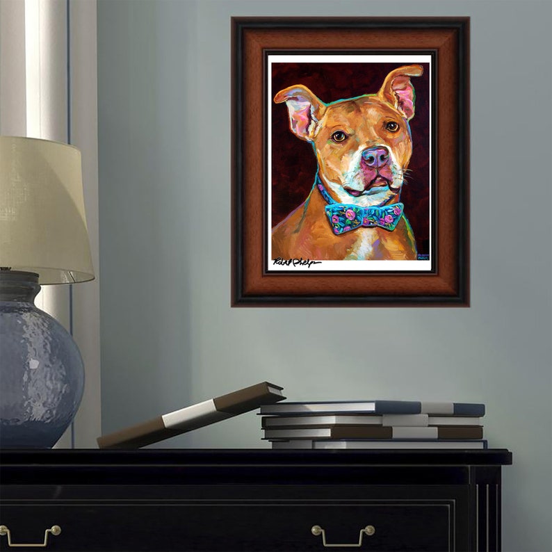 Puede incluir: Una pintura enmarcada de un perro marr&oacute;n y blanco con una pajarita azul con flores rosas. El perro est&aacute; mirando directamente al espectador. La pintura est&aacute; sobre un fondo rojo oscuro.