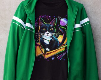 Puede incluir: Un gato blanco y negro con un collar dorado está en una caja con un fondo de galaxia. El gato lleva una chaqueta verde con rayas blancas.