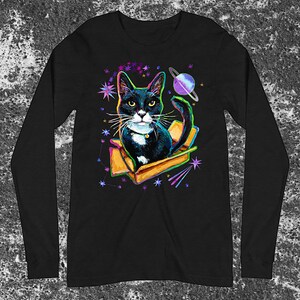 May include: A black and white cat with green eyes sits in a cardboard box against a black background with a galaxy print. The cat is wearing a gold collar and has a white chest. The galaxy print includes stars, a planet, and shooting stars.