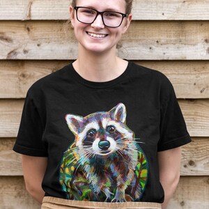 May include: A black t-shirt with a colorful illustration of a raccoon. The raccoon is looking directly at the viewer with a curious expression. The illustration is surrounded by green foliage.