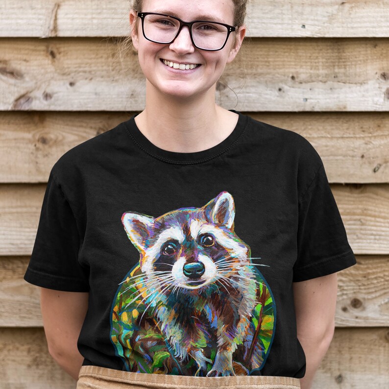 May include: A black t-shirt with a colorful illustration of a raccoon. The raccoon is looking directly at the viewer with a curious expression. The illustration is surrounded by green foliage.