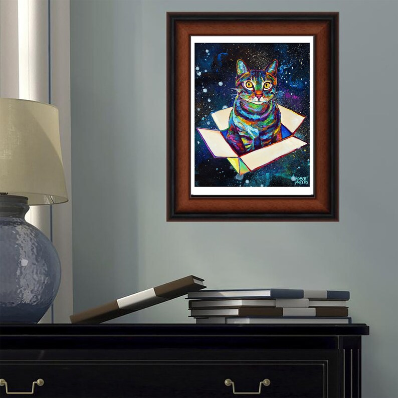 May include: A colorful painting of a cat sitting in a box against a dark blue background with white stars. The cat has green eyes and a striped pattern. The box is white with a yellow interior. The painting is framed in a brown wood frame.