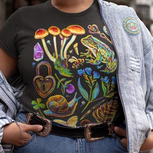 Puede incluir: Camiseta negra con un estampado gr&aacute;fico colorido que presenta setas, una rana, un caracol, un candado en forma de coraz&oacute;n, un cristal, una mariposa y flores.