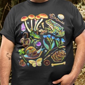 Puede incluir: Camiseta negra con una ilustraci&oacute;n colorida de setas, una rana, un caracol, una mariposa, una pi&ntilde;a, una llave, una cerradura y otros elementos inspirados en la naturaleza.