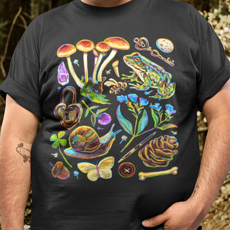 Puede incluir: Camiseta negra con una ilustraci&oacute;n colorida de setas, una rana, un caracol, una mariposa, una pi&ntilde;a, una llave, una cerradura y otros elementos inspirados en la naturaleza.