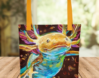 May include: A colourful tote bag featuring a whimsical painting of an axolotl, a type of salamander, with bright blue, yellow, and pink accents. The axolotl is depicted against a dark brown background with scattered colourful dots.