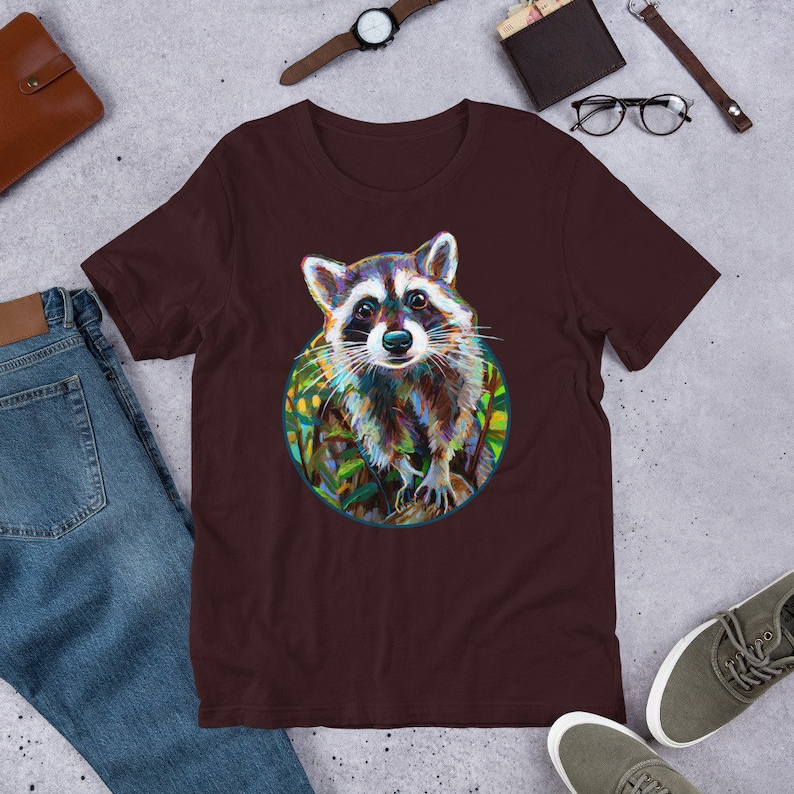 May include: A brown t-shirt with a colorful illustration of a raccoon in a circle. The raccoon is looking directly at the viewer with a curious expression. The background of the illustration is a green and yellow leafy pattern.