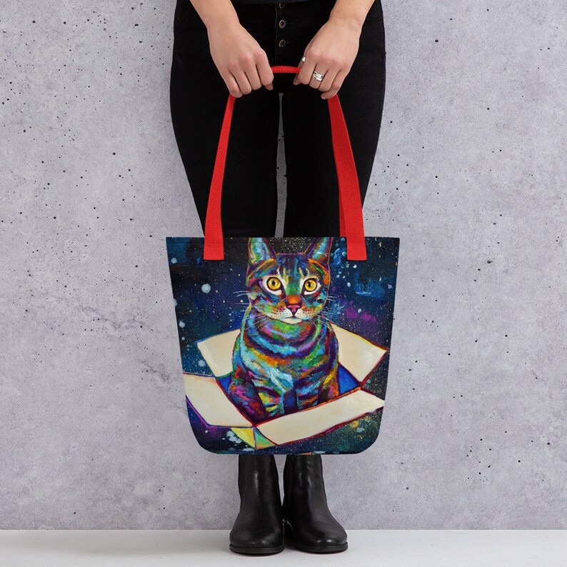 May include: A tote bag with red handles features a colorful cat sitting in a box against a dark blue and purple galaxy background.