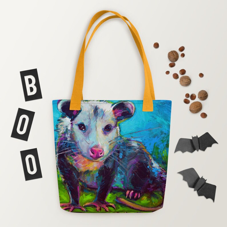 May include: A tote bag with a colorful painting of a possum on a blue background. The possum has a white face with black eyes and a pink nose. The bag has yellow straps.
