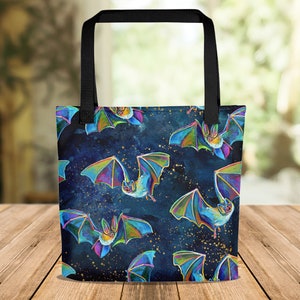 Könnte beinhalten: Eine schwarze Tragetasche mit einem bunten Fledermausmuster auf dunkelblauem Hintergrund. Die Fledermäuse sind schillernd und haben ein galaxieartiges Design. Die Tasche hat schwarze Henkel.