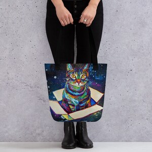 May include: A tote bag with a colorful cat illustration. The cat is sitting in a box with a galaxy background. The cat has yellow eyes and a white chest.