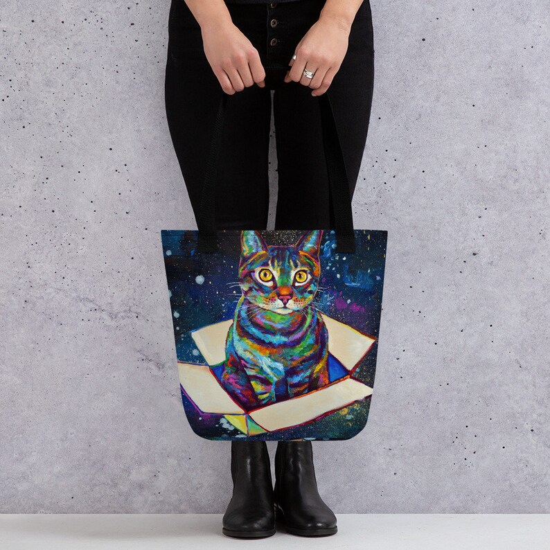 May include: A tote bag with a colorful cat illustration. The cat is sitting in a box with a galaxy background. The cat has yellow eyes and a white chest.