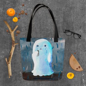 May include: A blue tote bag featuring a sad ghost illustration with tears, set against a rainy backdrop. The ghost is light blue with a white tissue. The bag has black handles.