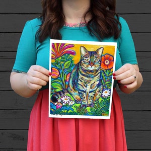 May include: A colorful painting of a tabby cat sitting in a garden with bright flowers and green foliage. The cat has green eyes and a white patch on its chest. The background is a bright yellow.