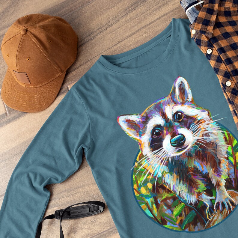 May include: A teal long-sleeve shirt featuring a colorful raccoon illustration. A brown baseball cap is visible to the left. A black camera strap and a plaid shirt are also in the image. The raccoon art has vibrant colors.