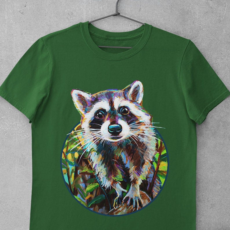 May include: Green t-shirt with a colorful illustration of a raccoon in a circle. The raccoon is looking at the viewer with a curious expression.