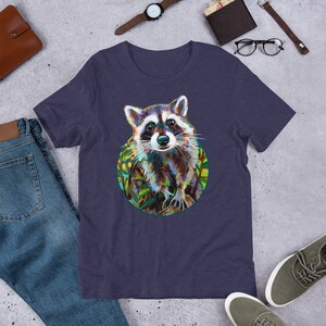May include: A navy blue t-shirt with a colorful illustration of a raccoon in a circle. The raccoon is looking directly at the viewer with a curious expression. The background of the illustration is a green and yellow leafy pattern.